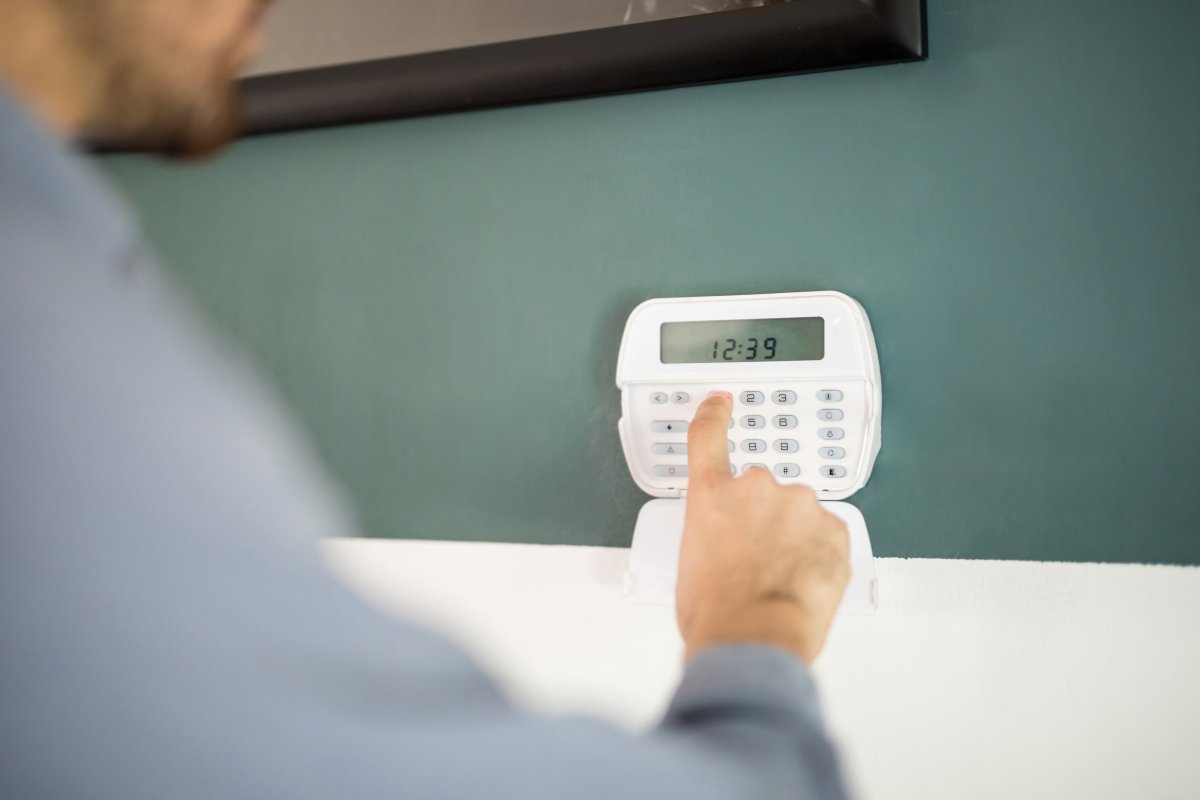 A person entering a code on a white wall-mounted security alarm keypad to arm their home security system.